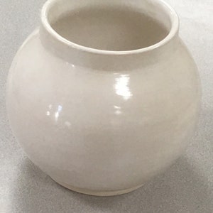 Może przedstawiać: Biały ceramiczny wazon o okrągłym korpusie i lekko rozszerzającym się otworze. Wazon ma gładkie, błyszczące wykończenie.