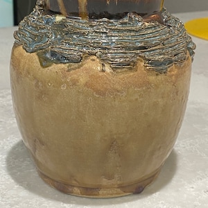 Puede incluir: Un jarrón de cerámica con un esmalte texturizado marrón y azul. El jarrón tiene un cuerpo redondeado y una abertura ancha.