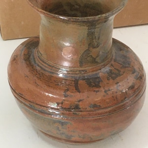 Puede incluir: Un jarrón de cerámica marrón con una base ancha y un cuello estrecho. El jarrón tiene un esmalte moteado con tonos de marrón, verde y negro.