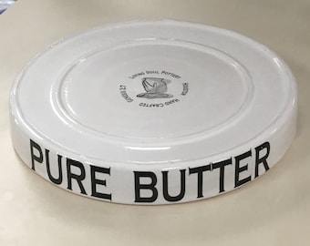 May include: A round white ceramic butter dish lid with a glossy finish. The side features the black text PURE BUTTER. The top center displays a black logo with the text LIVING SOUL POTTERY, HOUSTON, HAND CRAFTED, and the scripture reference GENESIS 2:7.