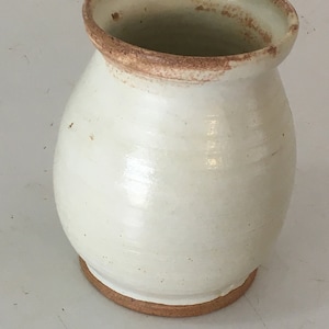 May include: A handmade, off-white ceramic vase with a rounded body and a slightly flared rim. The rim and base feature a natural, earthy brown color. The vase has a smooth, glossy finish and appears to be hand-thrown.