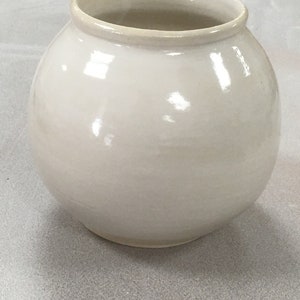 Może przedstawiać: Biały ceramiczny wazon o okrągłym korpusie i lekko rozszerzającym się rancie. Wazon ma błyszczące wykończenie.