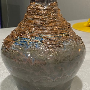 Puede incluir: Un jarrón de cerámica marrón y azul con una superficie texturizada y en capas. El jarrón tiene una base ancha y un cuello estrecho.