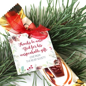May include: A white gift tag with a watercolor design of pine branches and berries. The tag has the text "Thanks be unto God for his unspeakable gift. Love from The Relief Society."  The tag is tied to a chocolate bar wrapped in gold foil and red ribbon.
