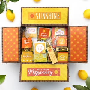May include: A cardboard gift box filled with various items, featuring yellow and orange sun-themed labels. The box flaps display sun patterns and text including "SUNSHINE" and "for a bright Missionary." Several small cards with inspirational messages are included.