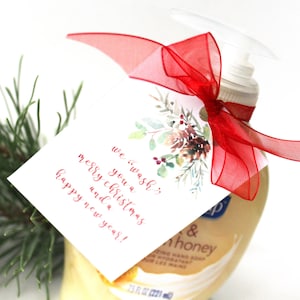 Op de afbeelding: Een wit cadeau-etiket met een aquarelontwerp van dennentakken, bessen en een dennenappel. Het etiket heeft de tekst "we 'wash' you a merry christmas and a happy new year!" in rood gedrukt. Het etiket is met een rood lint aan een fles handzeep gebonden.