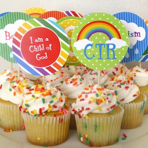 May include: A plate of cupcakes decorated with colorful sprinkles and topped with round paper circles with rainbow designs and religious messages. The messages include "I am a Child of God", "CTR", and "Baptism".