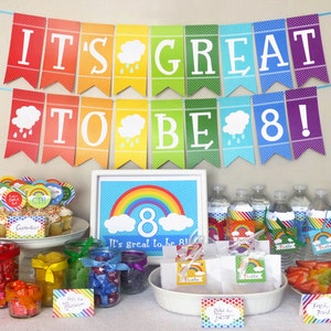 May include: Rainbow themed party decorations for an 8th birthday. The decorations include a banner that says "It's great to be 8!" and cupcakes with rainbow sprinkles. There are also rainbow-colored candies in jars and a rainbow-colored fruit platter.