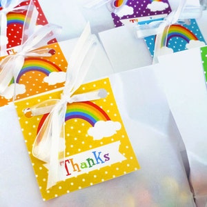 May include: A set of small white favor bags with rainbow and cloud designs. Each bag has a yellow tag with white polka dots and a rainbow with the word "Thanks" printed on it.