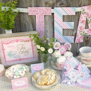 Könnte beinhalten: Eine rosa und blaue Tee-Party-Tischdekoration mit Cupcakes, Keksen und Teetassen. Der Tisch ist mit einem Banner mit der Aufschrift "TEA" und einem Schild mit der Aufschrift "Willkommen zur Tee-Party" dekoriert.