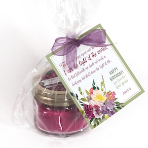 Puede incluir: Una pequeña vela de vidrio con una vela rosa en su interior. La vela está envuelta en celofán transparente y atada con una cinta morada. Una etiqueta de regalo verde y blanca con diseños florales y el texto "The spoke as again ants th I am the light of the world. he that followeth me shall not walk in darkness, but shall have the light of life. HAPPY BIRTHDAY! Love from the Relief Socialy JOHN 8-12" está unida al celofán.