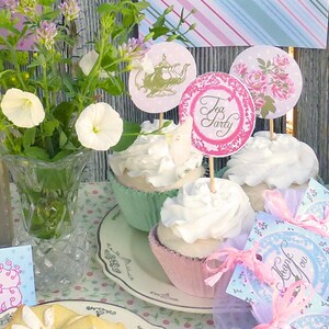 Puede incluir: Una mesa preparada para una fiesta de té con cupcakes, tazas de té y favores. Los cupcakes están decorados con glaseado blanco y adornados con flores rosas y blancas. Los favores son pequeñas bolsas con cintas rosas y azules y el texto "Gracias".