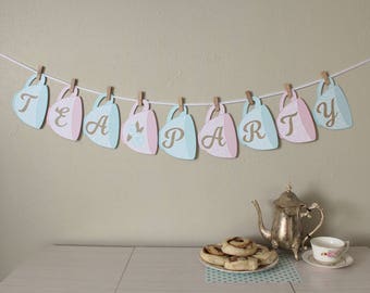 May include: A tea party banner featuring light blue and pink teacups with the letters "TEA PARTY" on them. The banner is strung with twine and hung on a wall. There are also pastries and a silver teapot on a table.