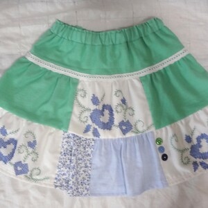 May include: A tiered skirt featuring a mint green elastic waistband and upper section, accented with white panels displaying blue cross-stitched floral heart patterns, light blue fabric segments, a small floral print panel, and three small round buttons.