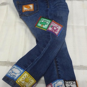 Peut inclure: Jean bleu pour enfant avec des écussons colorés. Les écussons comportent les mots "orange", "vert", "rose", "bleu", "jaune" et "noir", ainsi que des illustrations de glace, d'arbre, de fleur, de nuage, de soleil et de chat.