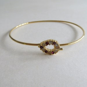 May include: A gold bangle bracelet with a small, round, jeweled clasp. The clasp is made of gold and has a circle of clear and red rhinestones.