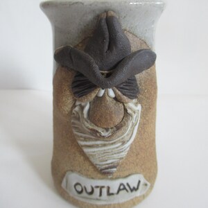May include: A brown ceramic mug with a sculpted face and a cowboy hat. The mug has a white glaze and the word "OUTLAW" is written on the front.