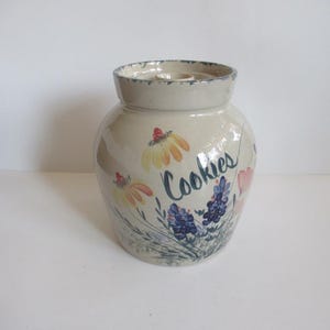 May include: A white ceramic cookie jar with a floral design and the word "Cookies" written on it.