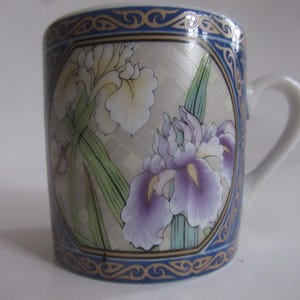 May include: A ceramic mug with a white handle and a floral design. The mug features a blue border with gold accents and a central panel with iris flowers in shades of purple, white, and green. The mug is approximately 4 inches tall.