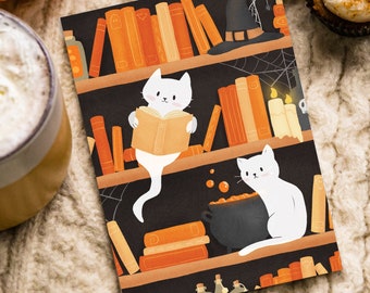 Può includere: Un gatto bianco che legge un libro su uno scaffale pieno di libri. Lo scaffale è decorato con una ragnatela e candele. C'è un secondo gatto bianco seduto sullo scaffale accanto a un calderone.