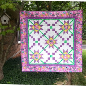 Op de afbeelding: Een paarse en groene quilt met een wit centrum en een roze bloemenrand. De quilt hangt aan een waslijn buiten.
