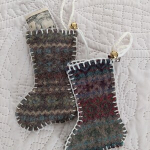 May include: Two small, hand-sewn stockings made from repurposed wool sweaters. The stockings are decorated with white stitching and have a bell attached to the top. One stocking has a dollar bill tucked inside.
