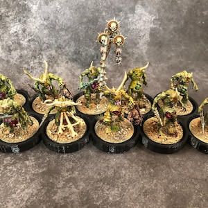 Peut inclure: Un ensemble de 10 figurines miniatures peintes en vert et blanc avec des cornes et des armes, pos&eacute;es sur des socles noirs. Les figurines sont dispos&eacute;es en cercle autour d'une figurine centrale avec un b&acirc;ton surmont&eacute; d'un cr&acirc;ne.