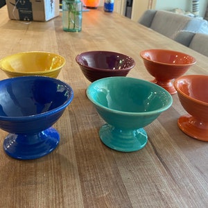 May include: Six colourful ceramic pedestal bowls in various shades of blue, green, yellow, orange, and red. The bowls are arranged on a wooden table.