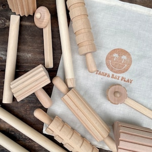 May include: A collection of unfinished wooden play tools, including rolling pins, a mallet, and a textured block, arranged on a wooden surface. A canvas bag with a smiling sun logo and the text "TAMPA BAY PLAY" is also present.