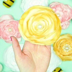 May include: A large, yellow fabric flower with a pearl center is held in a hand. Other fabric flowers in pink and yellow, along with white yarn and felt bees, are scattered on a light green surface. The yellow flower is the focal point.