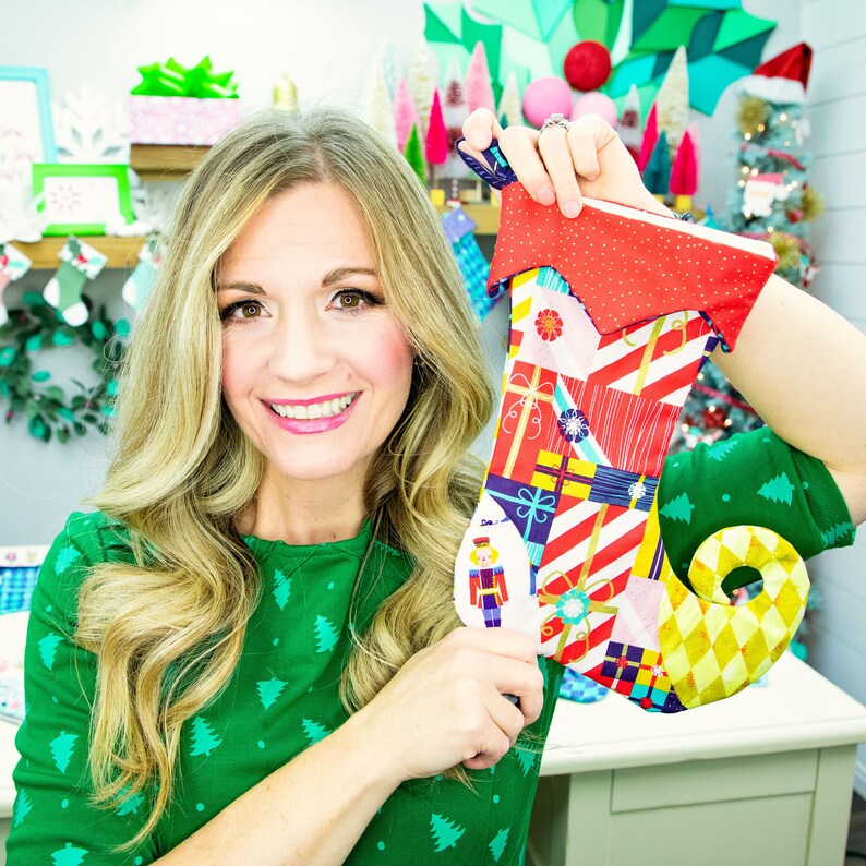May include: A woman is holding up a colourful Christmas stocking with a pattern of presents and a nutcracker design. The stocking has a red cuff and a yellow and gold toe.