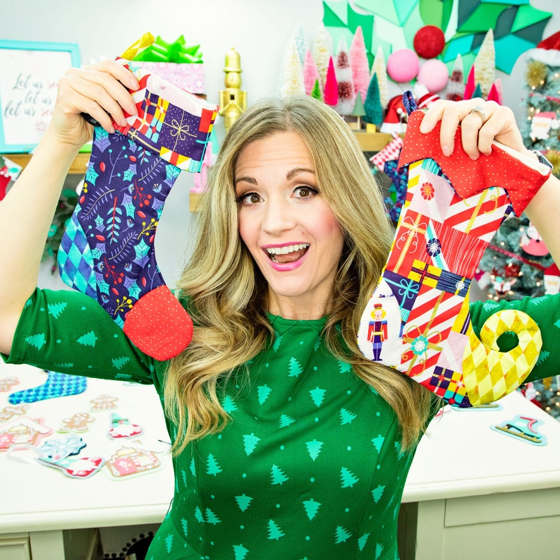 May include: A woman wearing a green dress with a Christmas tree pattern is holding two Christmas stockings. One stocking is blue with a white and green pattern and the other is red, white, and yellow with a nutcracker design.