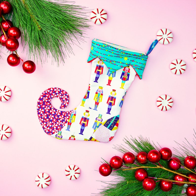 May include: A white Christmas stocking with a colourful nutcracker pattern. The stocking has a pointed toe and a green and blue cuff. The stocking is surrounded by red and white candy canes and a green pine branch with red berries.