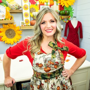 Puede incluir: Una mujer con una camisa roja y un delantal blanco con estampado de otoño. El delantal tiene un ribete verde y marrón y un lazo atado alrededor del cuello. El delantal presenta una cornucopia, calabazas y otras imágenes de la cosecha de otoño.