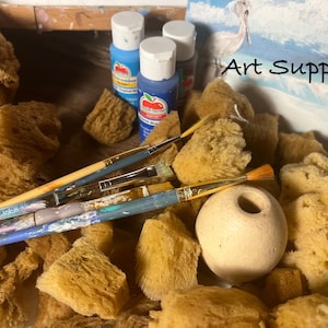 May include: An assortment of art supplies, including paintbrushes, natural sponges, and paint bottles. The image features the text "Art Supplies" in black. The color palette is dominated by shades of blue, yellow, and beige.