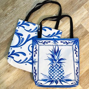May include: Two white tote bags with black handles. One bag features a blue floral design, while the other showcases a blue pineapple illustration within a blue border. The bags are made of fabric.