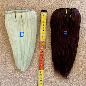 May include: Two bundles of hair extensions, one light green and one dark burgundy, are displayed next to a ruler. The ruler shows measurements in inches. The light green bundle is labeled "D" and the burgundy bundle is labeled "E".
