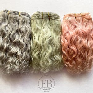 May include: Three bundles of wavy hair extensions in shades of gray, light green, and pink. Each bundle is secured with a thin, gold-colored band. The hair has a natural, textured appearance, suggesting a focus on hair styling and beauty products.