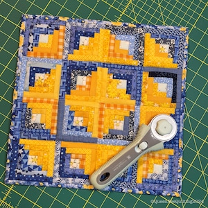 Könnte beinhalten: Ein blauer und gelber Quiltblock mit einem Blockhausmuster. Der Quiltblock ist aus verschiedenen Stoffen gefertigt, darunter Blumenmuster, Gingham und einfarbige Stoffe. Ein Rollschneider liegt auf dem Quiltblock.