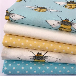 Könnte beinhalten: Ein Stapel Stoffmuster in verschiedenen Mustern und Farben. Der oberste Stoff ist hellblau mit einem Bienenmuster. Darunter befindet sich ein weißer Stoff, dann ein gelber Stoff mit weißen Punkten, ein weiterer weißer Stoff mit einem Bienenmuster und schließlich ein blauer Stoff mit weißen Punkten.