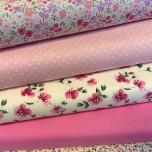 May include: Several fabric bolts with floral and polka dot designs. The top layer displays a white background with pink and purple flowers. Beneath is a pink polka dot fabric, followed by a floral print with pink flowers, and a solid pink fabric.