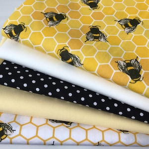 May include: A stack of five fabric squares with different patterns. The top square is yellow with a honeycomb pattern and black bees. The second square is white. The third square is black with white polka dots. The fourth square is a solid yellow. The bottom square is white with a honeycomb pattern and black bees.