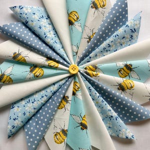 May include: A circular display of fabric triangles in various patterns, including white, light blue with white polka dots, and light blue with white floral and bee designs. A yellow button is at the centre.