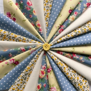 May include: A circular design composed of fabric triangles in a variety of patterns and colours. The fabrics feature floral prints, polka dots, and solid colours in shades of blue, yellow, and white. A yellow button is at the centre, securing the triangles.