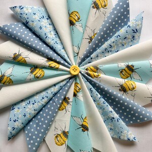 Könnte beinhalten: Eine kreisförmige Anordnung von Stoffdreiecken mit Bienen- und Blumenmustern in Weiß, Hellblau und Marineblau. Ein gelber Knopf sichert die Mitte. Die Stoffmuster umfassen Bienen, Tupfen und kleine Blumen.