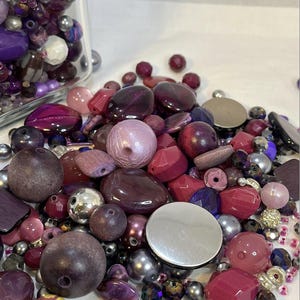 May include: A selection of purple and silver beads in diverse forms. The beads include spherical, faceted, and flat disc shapes, with both matte and glossy finishes. Some beads are contained in a clear glass jar.