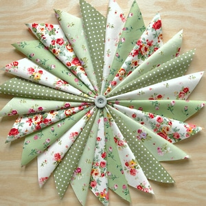 May include: A green and white fabric starburst with a floral pattern. The fabric is arranged in a circle with the points of the starburst radiating outwards. The centre of the starburst is a small button.
