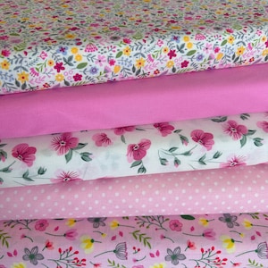 May include: A stack of five folded fabric bolts featuring various patterns. The top layer has a dense floral print, followed by a solid bright pink, a white fabric with large pink flowers, a light pink with white polka dots, and a floral print base.