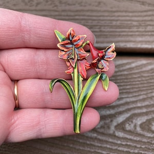 Könnte beinhalten: Blumenbrosche mit einem stilisierten Amaryllis-Design. Die Brosche hat rote und orangefarbene Blütenblätter mit goldenen Akzenten, grüne Blätter und einen grünen Stiel. Die Brosche ist aus Metall und Emaille gefertigt.