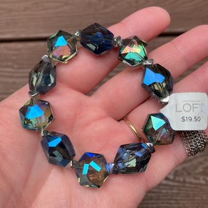 May include: A silver bracelet with iridescent blue and green faceted glass beads. The bracelet is on a hand and has a price tag that reads "LOFT $19.50".